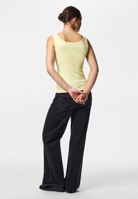 Femme aux cheveux foncés attachés en chignon, portant un haut sans manches jaune clair et un jean noir à jambes larges, debout avec les mains jointes dans le dos.