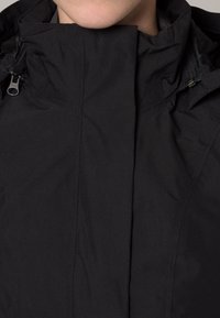 Veste isolante noire avec un col haut, des poches zippées, une texture de tissu douce et des éléments de design minimalistes.