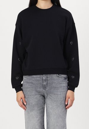 Mujer con sudadera negra de manga larga con pequeños diseños de corazones blancos en las mangas y vaqueros grises desgastados.