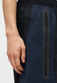 Κοντινό πλάνο ενός χεριού δίπλα σε σκούρο μπλε και μαύρο σορτς με κάθετη τσέπη με φερμουάρ και διπλούς μαύρους μηχανισμούς φερμουάρ.