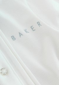 Tessuto bianco con trama a griglia sottile. Marchio "BAKER" in grigio chiaro, accanto a un'etichetta circolare con la scritta "BAKER BY RED BAKER."