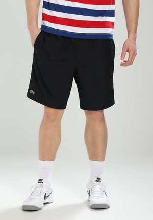 Homme portant un short noir avec un petit logo vert sur la jambe gauche, des chaussettes blanches Nike, des baskets blanches Nike, et un t-shirt rayé rouge, blanc et bleu.