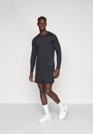 Fekete hosszú ujjú sportpóló hozzáillő rövidnadrággal, könnyű anyagból készült. Fehér sportcipő és bokazokni egészíti ki az öltözéket.