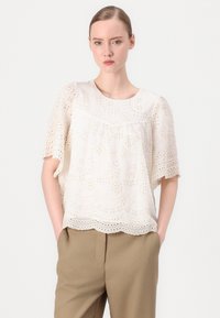 Vit spetsblus med korta ärmar, med blommönstrade utskärningar och vågig nederkant. Bärs över beige byxor.