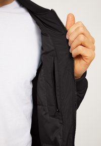 Black puffer jacket with ribbed cuffs and collar, featuring a zippered interior pocket. The fabric appears smooth and lightweight.