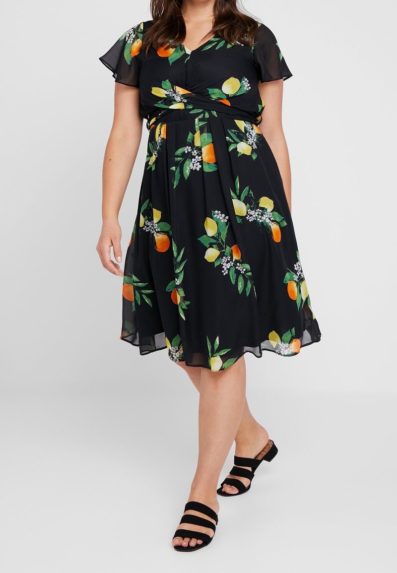 Sort blomsterkjole med citron- og appelsinmønstre, korte flæseærmer, tilpasset talje og flydende nederdel. Kombineret med sorte slip-in sandaler.