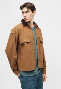 Junger Mann trägt eine braune Jacke mit Knöpfen über einem grünen Hemd und blauen Jeans, schaut nach links vor einem einfarbigen Hintergrund.