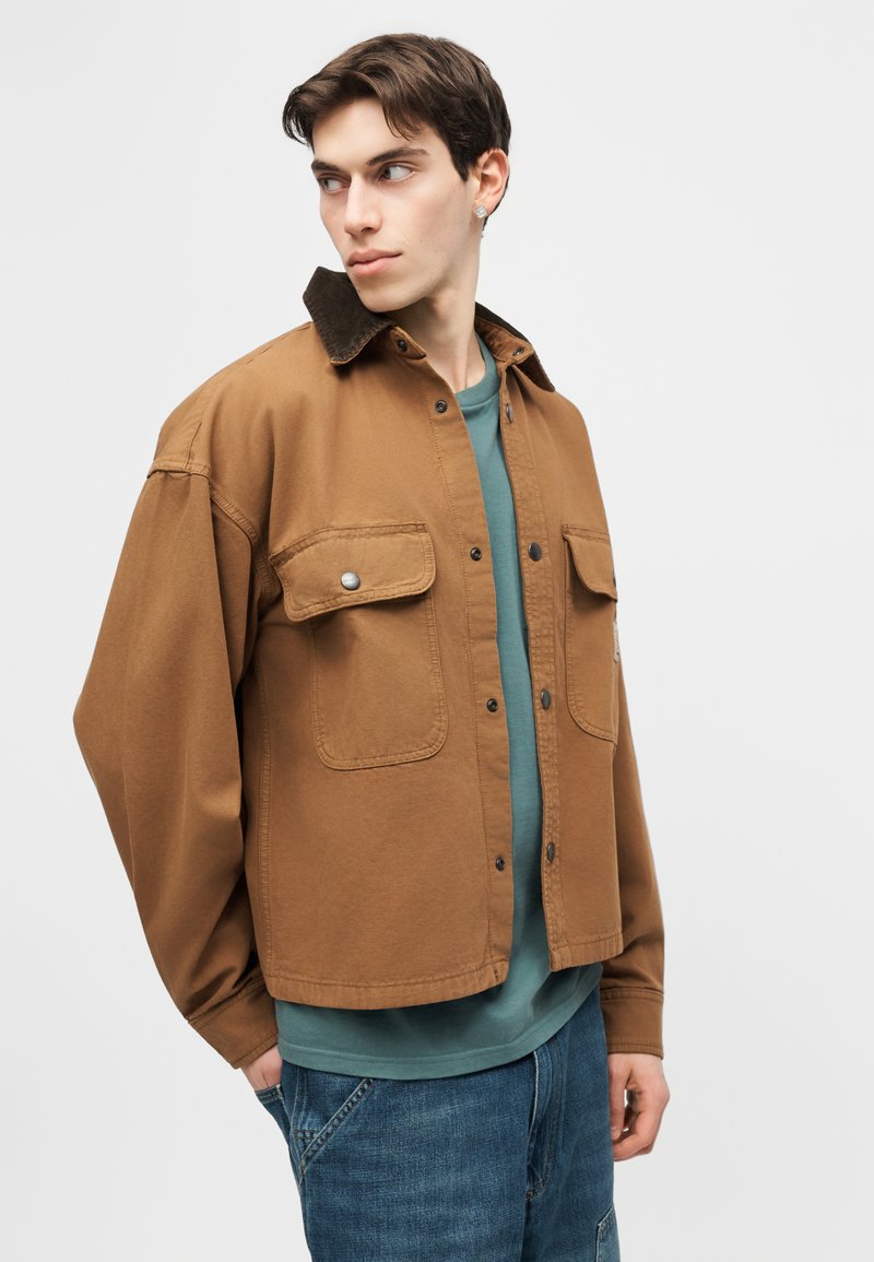 Junger Mann trägt eine braune Jacke mit Knöpfen über einem grünen Hemd und blauen Jeans, schaut nach links vor einem einfarbigen Hintergrund.