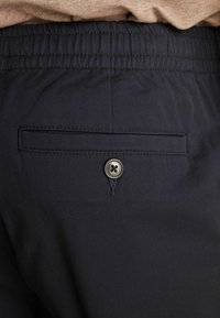 Tmavě modré kalhoty s texturovanou tkaninou, které mají elastický pas a zadní kapsu zabezpečenou kovovým knoflíkem.