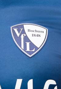 Λογότυπο VfL Bochum 1848 σε μπλε αθλητικό ύφασμα, με λευκό και ναυτικό ασπίδα που περιλαμβάνει τα αρχικά του συλλόγου και το έτος ίδρυσής του.