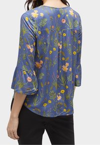 Blouse bleue à motifs floraux variés en rose, jaune et vert, avec manches évasées et coupe décontractée, confectionnée en tissu léger.