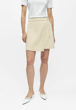 Femme portant une jupe portefeuille beige, un haut blanc à manches courtes et des chaussures blanches à bout pointu, debout devant un fond uni.