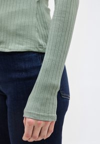 Κοντινό πλάνο ενός ατόμου που φοράει μακρυμάνικο, ριμπ πλεκτό ανοιχτοπράσινο τοπ και σκούρα μπλε τζιν παντελόνια, με το χέρι χαλαρό στο πλάι.