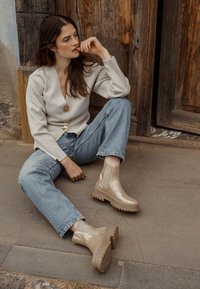 Botines gruesos en color marrón claro con acabado brillante, paneles laterales elásticos y suela resistente. El modelo lleva unos jeans de color azul claro y una blusa beige, sentado al aire libre.