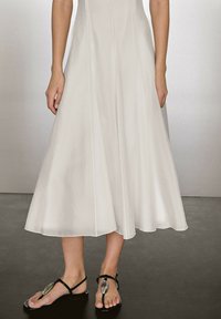 Weißes, schwingendes Midikleid mit vertikalen Nähten, das eine A-Linie-Silhouette hat. Schwarze Sandalen mit einem kreisförmigen Akzent runden das Outfit ab.