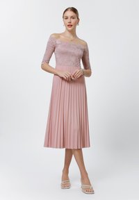 Off-the-shoulder pink dress with lace bodice, 3/4 sleeves, and pleated skirt, paired with white strappy sandals. Smooth texture throughout.