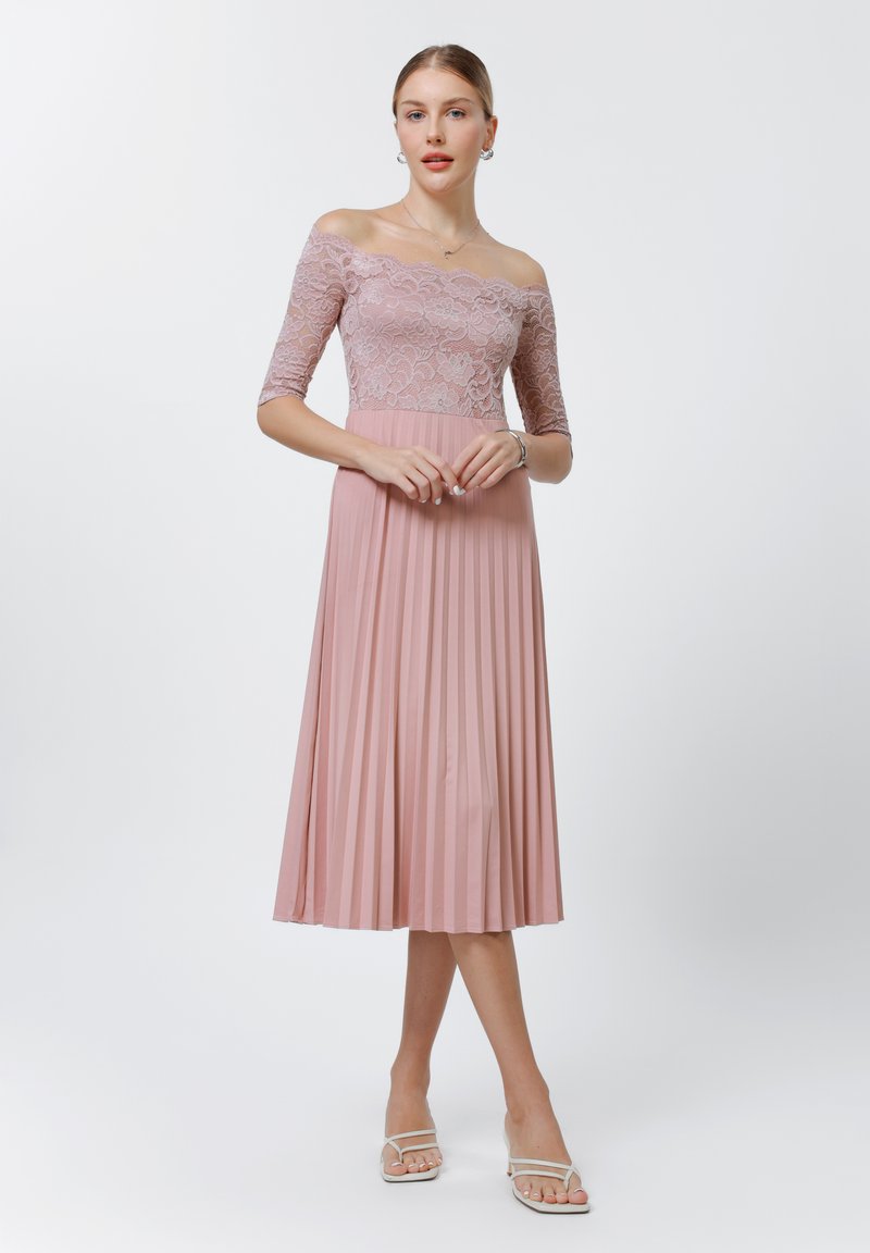 Robe rose décolletée avec corsage en dentelle, manches 3/4 et jupe plissée, associée à des sandales blanches à lanières. Texture lisse sur l'ensemble.