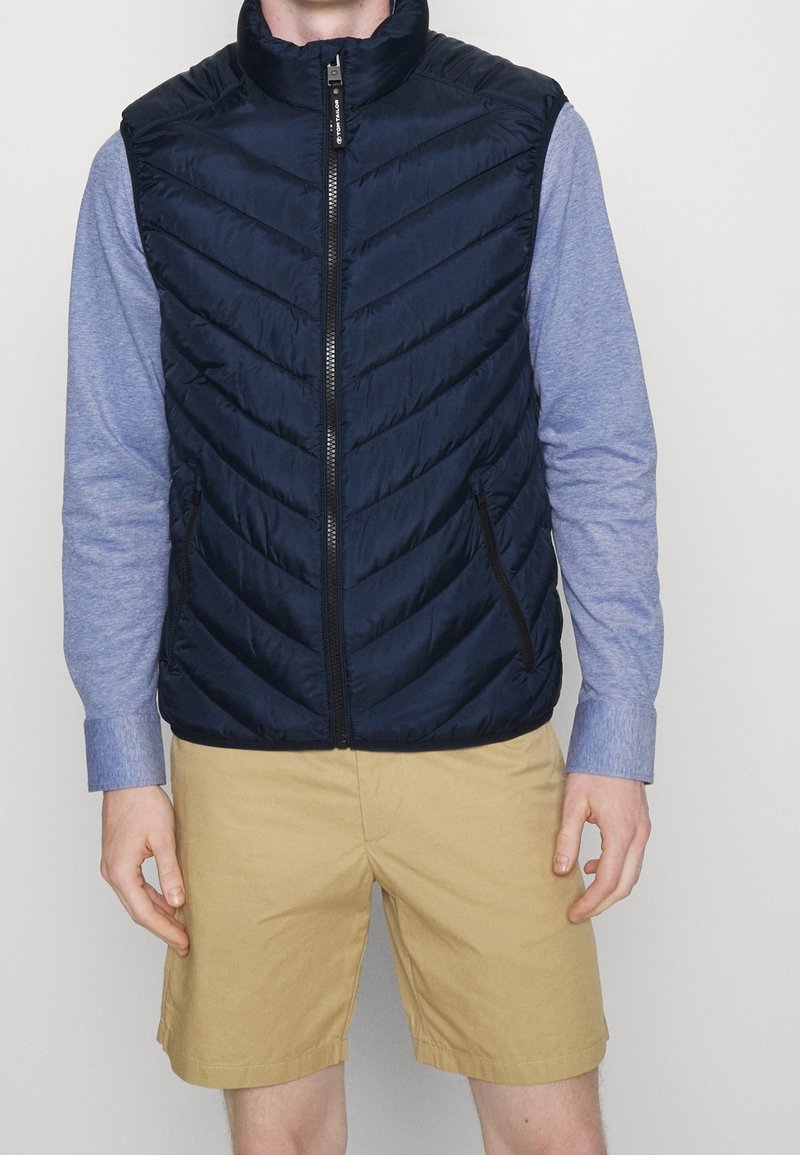 Homme portant un gilet matelassé bleu marine zippé sur une chemise à manches longues bleu clair avec des shorts beiges, debout devant un fond uni.