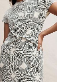 Schwarzes und weißes gemustertes Oberteil mit kurzen Ärmeln, ausgestattet mit geometrischen Designs und texturierter Stoffoberfläche, Knoten-Detail an der Taille, glatte Oberfläche.