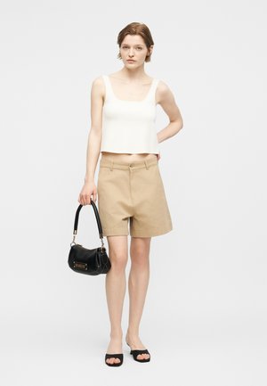 Femme portant un débardeur blanc court et un short beige, tenant un sac à main noir, debout avec une main sur la hanche contre un fond uni.