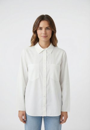 Giovane donna con capelli castani di media lunghezza che indossa una camicia bianca con bottoni e jeans azzurri, in piedi davanti a uno sfondo bianco uniforme.