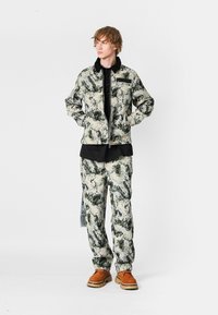 Conjunto de chaqueta y pantalones multicolores con patrones abstractos en verde y beige. La chaqueta cuenta con una cremallera, bolsillo en el pecho y cuello negro.