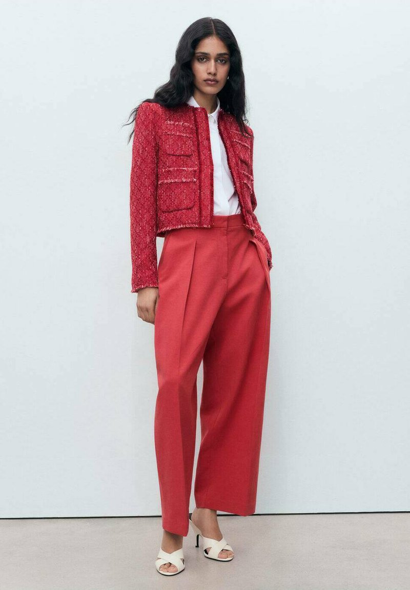 Rote Tweedjacke mit aufgesetzten Taschen, kombiniert mit lockeren roten Hosen. Das Modell trägt eine weiße Bluse und weiße Sandalen mit offenen Zehen.