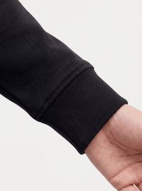 Primo piano del polso di una persona che indossa una manica lunga nera con polsino a coste su uno sfondo chiaro uniforme.