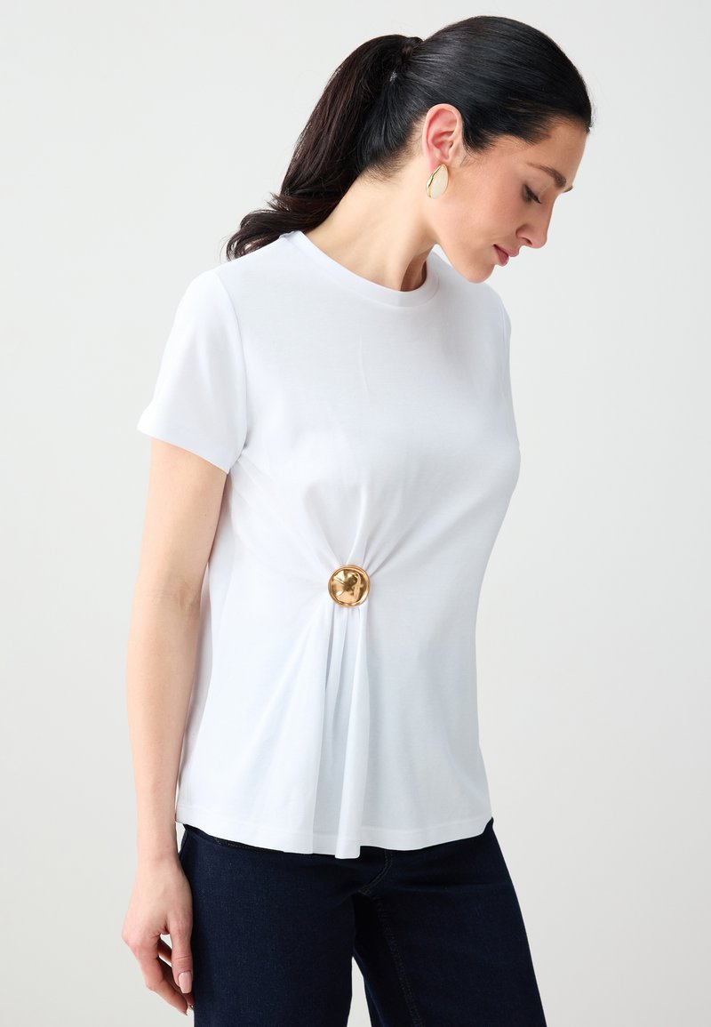 Femme aux cheveux foncés attachés en queue de cheval, portant un haut blanc à manches courtes avec un détail de boucle circulaire dorée à la taille, associée à un jean foncé.