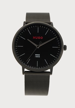 Montre-bracelet noire Hugo avec logo rouge, fines marques d'heure argentées, aiguilles fines argentées et bracelet en métal mesh, affichant l'heure 10:09:36.
