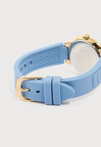 Μπλε λουράκι ρολογιού από σιλικόνη με χρυσό αγκράφα και κούμπωμα, υφή στο εσωτερικό του λουριού, προσαρτημένο σε ρολόι με χρυσή κάσα και ορατό πίσω καπάκι.