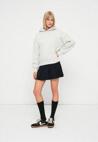 Felpa oversized grigia chiara con cappuccio regolabile; abbinata a una gonna plissettata nera, calze nere fino al ginocchio e sneakers nere con dettagli bianchi.