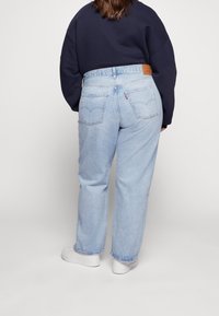 Osoba ubrana w jasnoniebieskie dżinsy z wysokim stanem i prostymi nogawkami, granatowy sweatshirt oraz białe sneakersy, stojąca plecami na jednolitym tle.