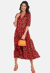 Robe midi florale avec un fond noir, ornée de coquelicots rouges et orange, à manches courtes bouffantes et jupe à volants. Les accessoires comprennent un sac à main orange.