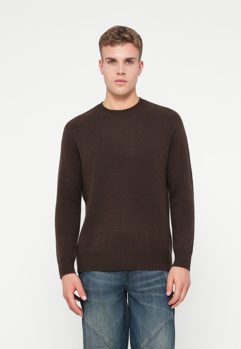 Ung man med kort hår som bär en mörkbrun polotröja och blå jeans, stående mot en enfärgad vit bakgrund.