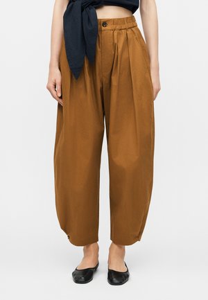 Καφέ παντελόνι με ψηλή μέση και φαρδιά πόδια, με κουμπί και ελαστική μέση, συνδυασμένο με μαύρα πλατιά παπούτσια και μαύρο κοντό τοπ δεμένο στη μέση.