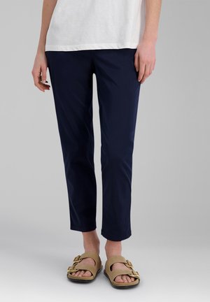 Person trägt marineblaue kurze Hosen, beige Sandalen mit Schnalle und ein weißes Oberteil vor einem einfarbigen hellgrauen Hintergrund.