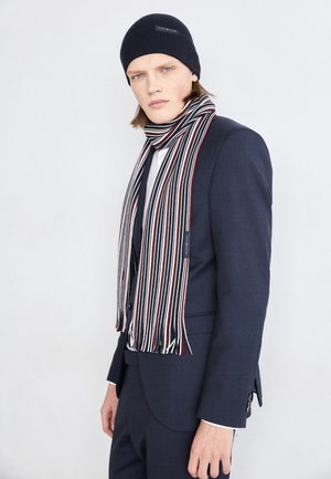 Junger Mann in marineblauem Anzug, schwarzer Mütze und gestreiftem rot-weiß-schwarzem Schal, stehend vor einem einfarbigen hellen Hintergrund.