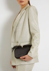 Sidabrinis blizgantis švarkas su struktūruotais pečiais, derinamas su juodu delniniu, turinčiu auksinį užsegimą ir plunksnų akcentus, laikomas rankoje.