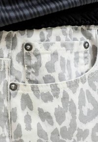 Gros plan d'une poche en denim clair avec un motif animalier gris et des rivets métalliques, superposée sous un tissu foncé rayé.