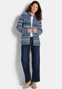 Strickjacke in verschiedenen Blautönen, mit hohem Kragen, Knopfverschluss und strukturiertem Stoff. Kombiniert mit dunkler Jeans und beigen Schuhen.