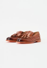Mocasines de cuero marrón con borlas, que presentan una textura suave, punta redondeada y suela naranja en contraste. Costuras detalladas a lo largo de los bordes.