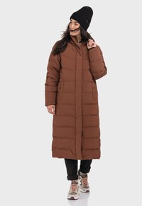 Brauner, langer Puffermantel mit einem Reißverschluss vorne, seitlichen Taschen und horizontalem Steppmuster; kombiniert mit schwarzen Hosen und beigen Sneakers.