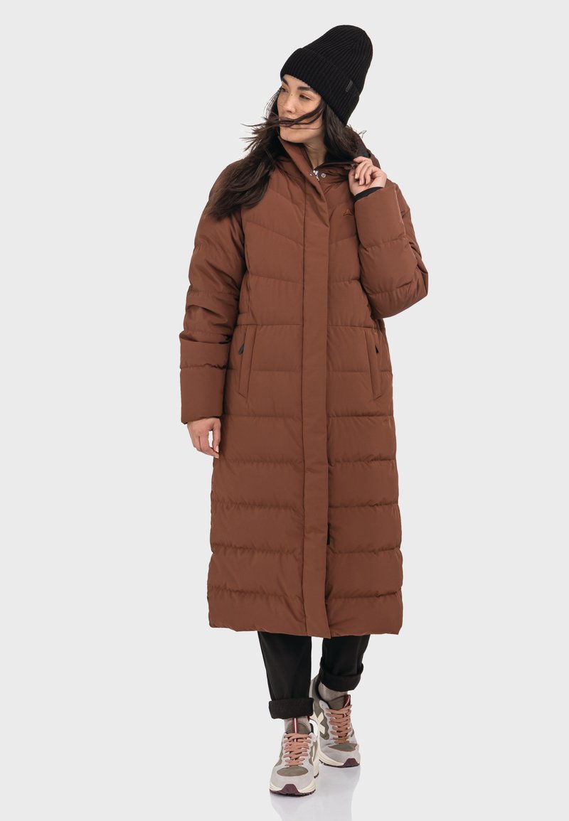 Brauner, langer Puffermantel mit einem Reißverschluss vorne, seitlichen Taschen und horizontalem Steppmuster; kombiniert mit schwarzen Hosen und beigen Sneakers.
