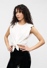 Λευκό crop top με κοντά μανίκια και δεσίματα στο μπροστινό μέρος, με μεταλλικές λεπτομέρειες στους ώμους. Συνδυάζεται με γυαλιστερό μαύρο κολάν.