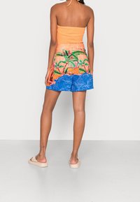 Femme portant un haut orange à dos nu, un short à imprimé tropical avec des palmiers et des vagues océaniques, et des sandales beiges à enfiler, se tenant de dos.