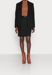 Svart kavaj över en brun ribbad polotröja, i kombination med en svart figursydd kjol och bruna anklarstövlar, allt mot en vit bakgrund.