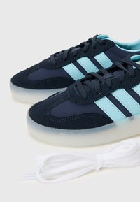 Sneakers de azul-marinho e azul claro com material em camurça e sintético, apresentando três riscas azuis, bico arredondado e atacadores brancos ao lado.