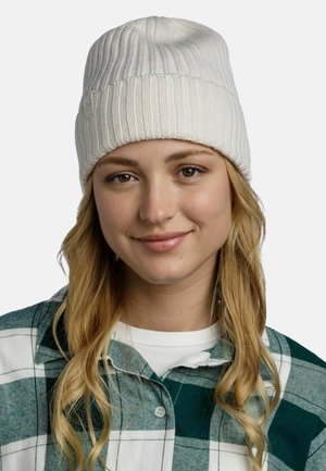 White knit beanie with ribbed texture. Worn slightly slouched, with a tag detail on the side. Model has long, wavy hair and a green plaid shirt.