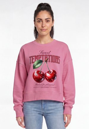 Femme aux cheveux foncés en queue de cheval portant un sweat rose avec de grosses cerises et le texte "Sweet Temptations" et un jean bleu clair.